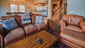 A spacious living area with a wooden staircase, leather sofas, a dining table, and a kitchen in the background. - Beautiful Custom Log Home - 1 Mile to Sunday River, Walk to Matterhorn (Newry)