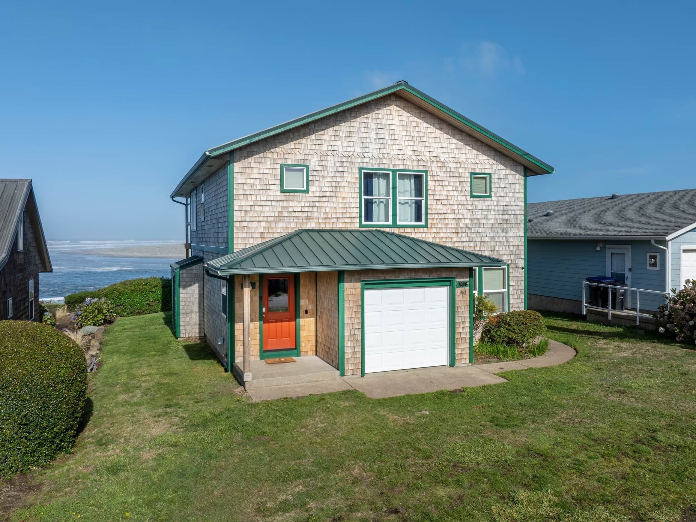 Une maison à deux étages, dotée d’un toit végétalisé et d’une porte de garage blanche, située près d’une plage et offrant une vue dégagée sur l’océan.