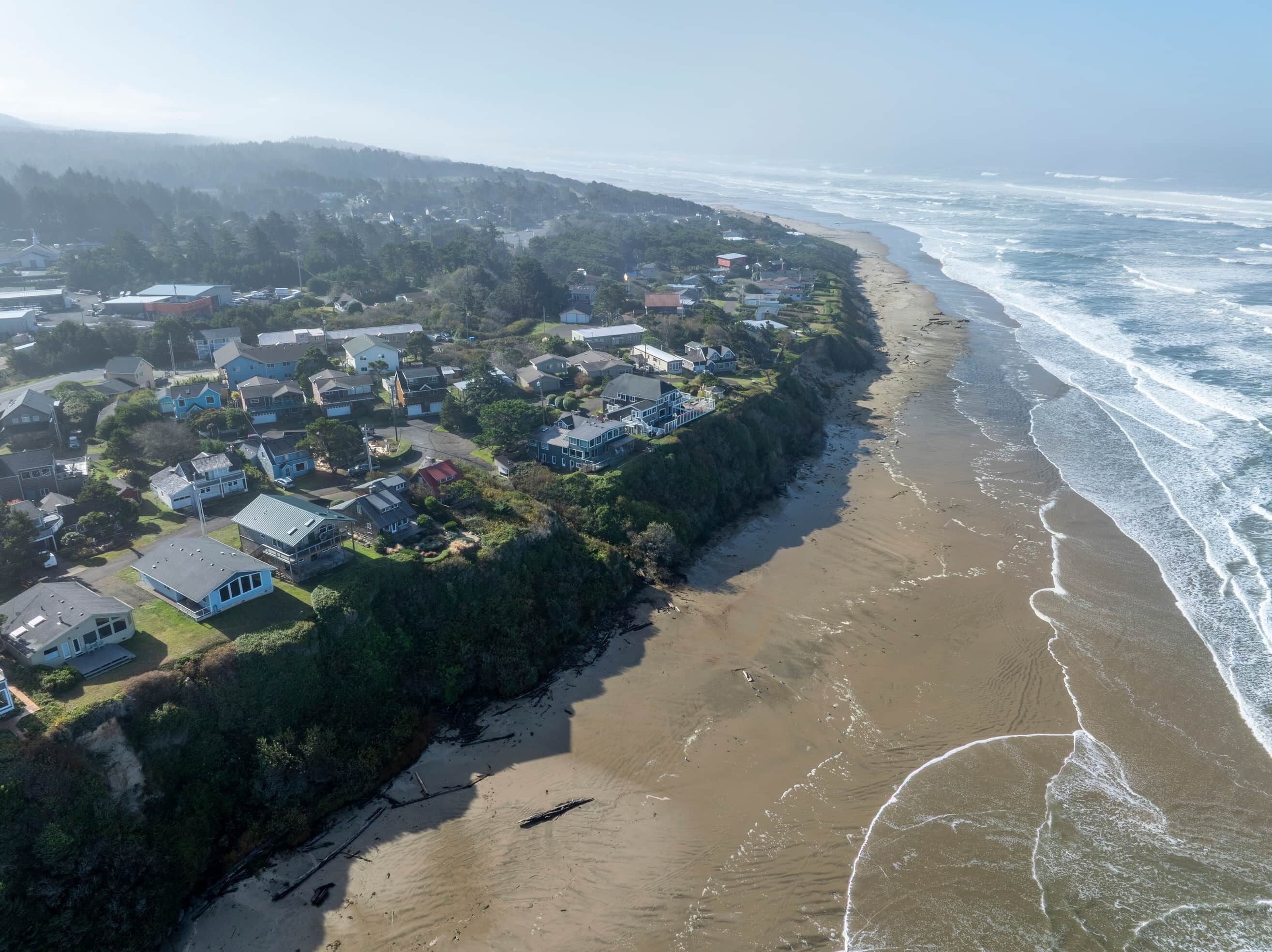 An aerial view of a coastal residential area with houses along the beach, clear skies, and waves approaching the shore.