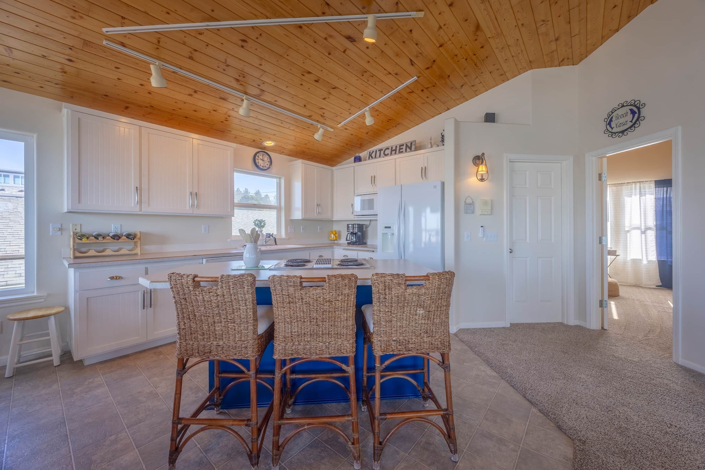 A modern kitchen with white cabinets, a central island with wicker chairs, and a clock on the wall.
