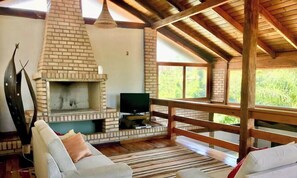 A rustic interior with a brick fireplace, a television, and a wooden deck overlooking greenery.