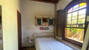 Quarto pequeno com cama de solteiro, teto de madeira, ventilador de teto, janela com vista para o exterior e uma mesa pequena com abajur.