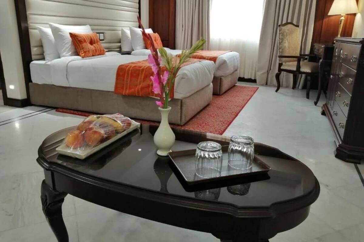 Een hotelkamer met een bed, een nachtkastje, een vaas met bloemen, een dienblad met fruit en glazen, en een dressoir.