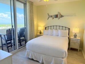 A hotel room with a bed, two bedside tables with lamps, a fish-shaped wall decoration, a balcony with chairs, and a view of the ocean.