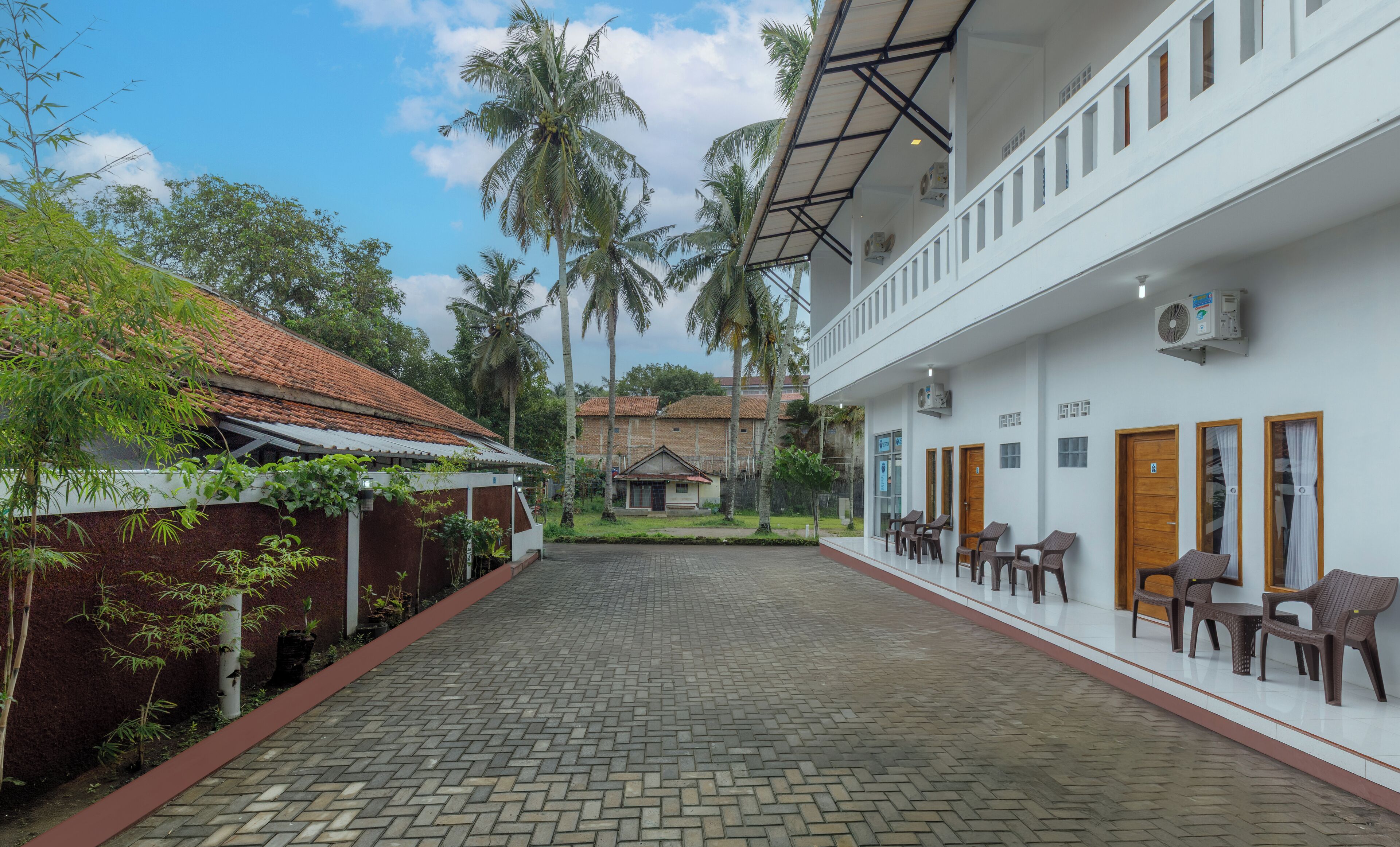 A courtyard with a paved walkway, a white building with multiple doors and windows, and palm trees in the background.