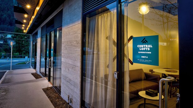 A modern hotel entrance with glass doors, a sign for Central Lofts Queenstown Unit 4, and a seating area with a table and chairs inside. - Central Lofts 4 & 5 - Accessible - Brand New! (Queenstown)