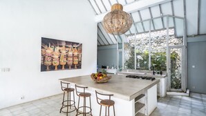 A kitchen with a large island, bar stools, a hanging light fixture, a stone wall, and a large window. - Villa Zen Bingin (Pecatu)
