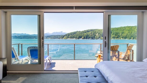 A hotel room with a large sliding glass door leading to a balcony with a view of a lake and mountains. There is a bed, a chair, and a small table on the balcony. - Hood Canal Resort in Union, WA (Union)
