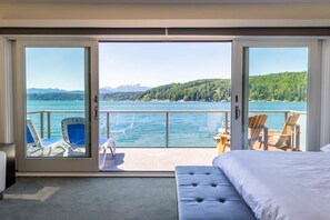 A hotel room with a large sliding glass door leading to a balcony with a view of a lake and mountains. There is a bed, a chair, and a small table on the balcony. - Hood Canal Resort in Union, WA (Union)