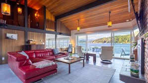 A spacious living area with a red leather sofa, a wooden coffee table, and a view of a lake through large windows. - Walls of Glass (Union)