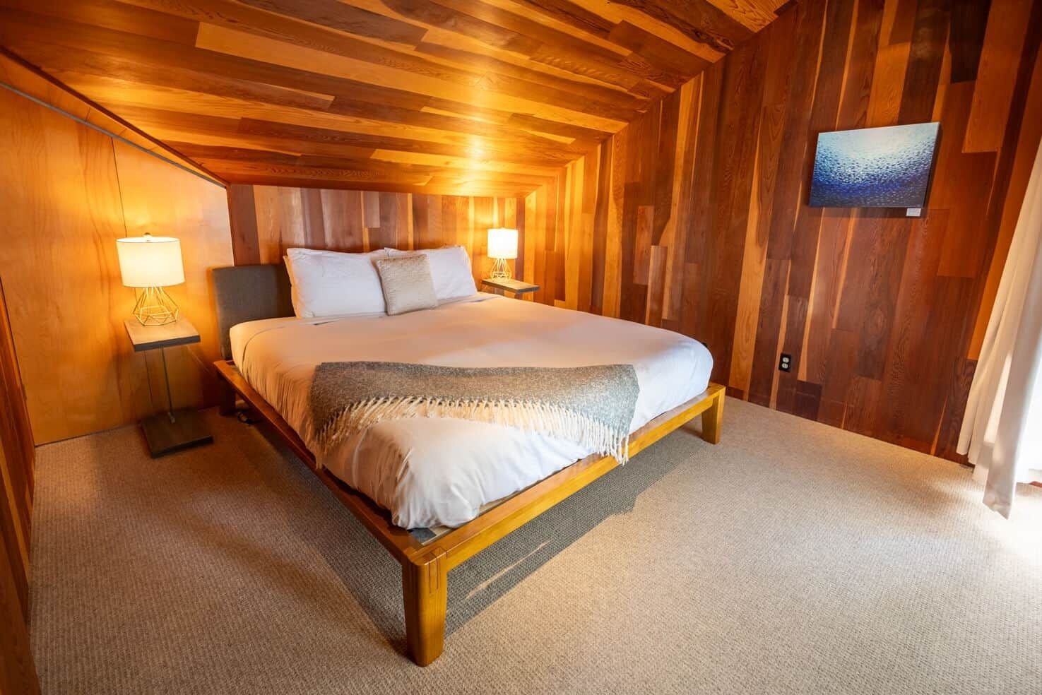 A wooden-paneled bedroom with a large bed, two bedside lamps, and a painting on the wall.