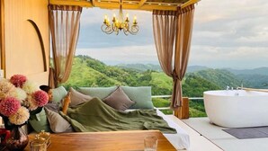 A bedroom with a bed, a large window, a chandelier, and a view of green hills. - Area21 Khaokho (Khao Kho)