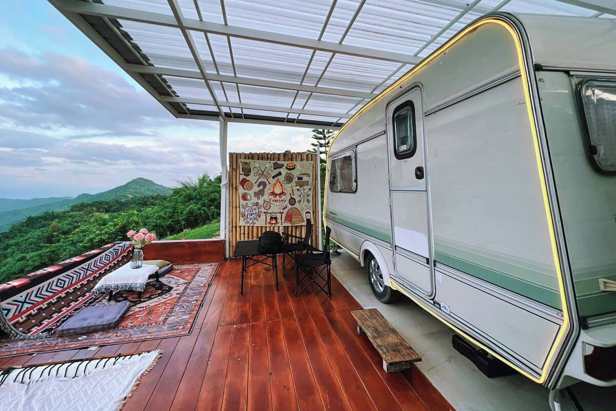 Eine überdachte Terrasse mit einem daneben stehenden Wohnmobil, mit Holzboden und Blick auf grüne Hügel.