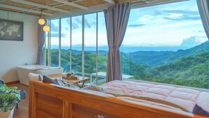 A bedroom with a large window, a wooden ceiling, a bed, a bedside table, and a view of a mountainous landscape. - Area21 Khaokho (Khao Kho)