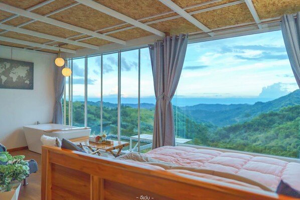 A bedroom with a large window, a wooden ceiling, a bed, a bedside table, and a view of a mountainous landscape. - Area21 Khaokho (Khao Kho)