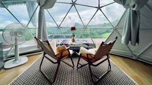 A room with a large window, a fan, a table with chairs, and a vase with a flower. - Area21 Khaokho (Khao Kho)