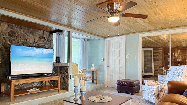 A living room with a stone wall, a television, a ceiling fan, and a view of the beach through a window. - Englewood Escape – Pet Friendly + Heated Pool (Englewood)