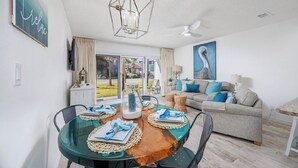 A dining area with a glass table, chairs, and a chandelier. There is a sofa and a painting in the background. - 🏖️ 5⭐️ Maravilla 4108!☀️Beautiful Remodel! 1 Bd 1.5 Bath w Bunks-Beach Svc 2Pools! (Miramar Beach)