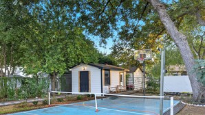 An outdoor basketball court with a net, surrounded by trees and a small house.