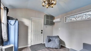 A room with a door, a blue suitcase, a gray bean bag chair, a gray couch, a ceiling light fixture, and a window with a view of bare trees.