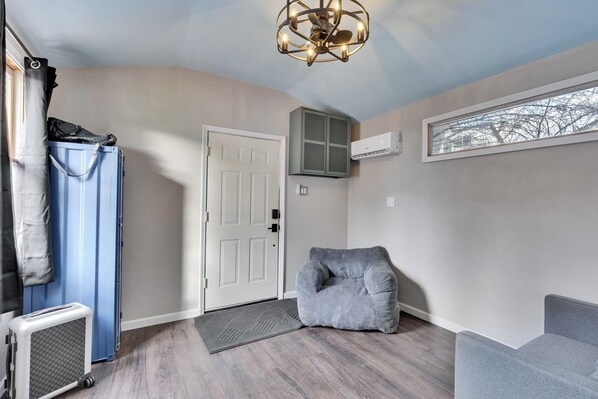 A room with a door, a blue suitcase, a gray bean bag chair, a gray couch, a ceiling light fixture, and a window with a view of bare trees.