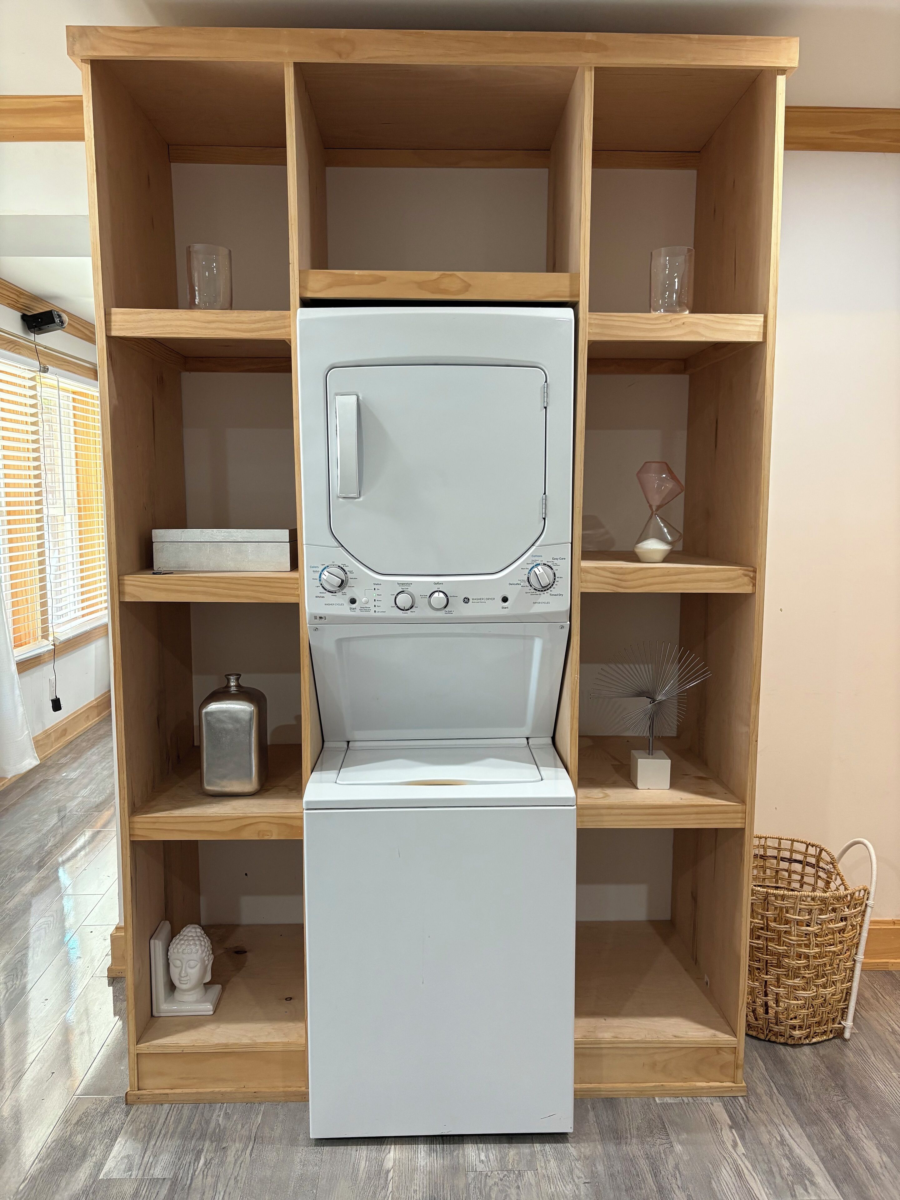 A white washing machine in a wooden cabinet with shelves, a small statue, and a basket.
