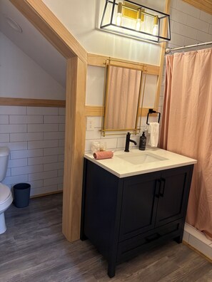 Un baño con un lavamanos blanco, un mueble negro, vigas de madera en el techo y un espejo con una lámpara sobre él.