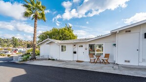 A single-story white mobile home with a garage, a small porch with two chairs, and a palm tree in the background.