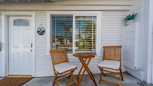A white house with a front door, a window with blinds, and two wooden chairs with cushions.