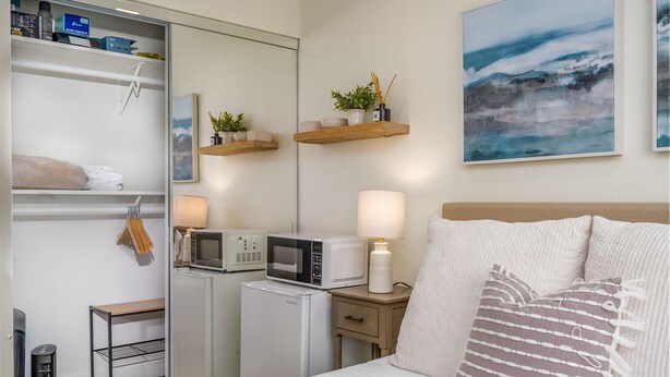 A neatly organized hotel room with a bed, a microwave, a refrigerator, a small table, and a shelf with decorative items.