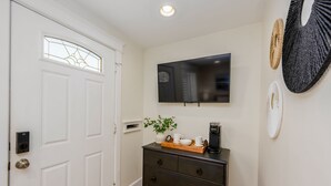 A hallway with a white door, a black coffee maker on a wooden tray, a white vase with a plant, and a mounted flat-screen TV.