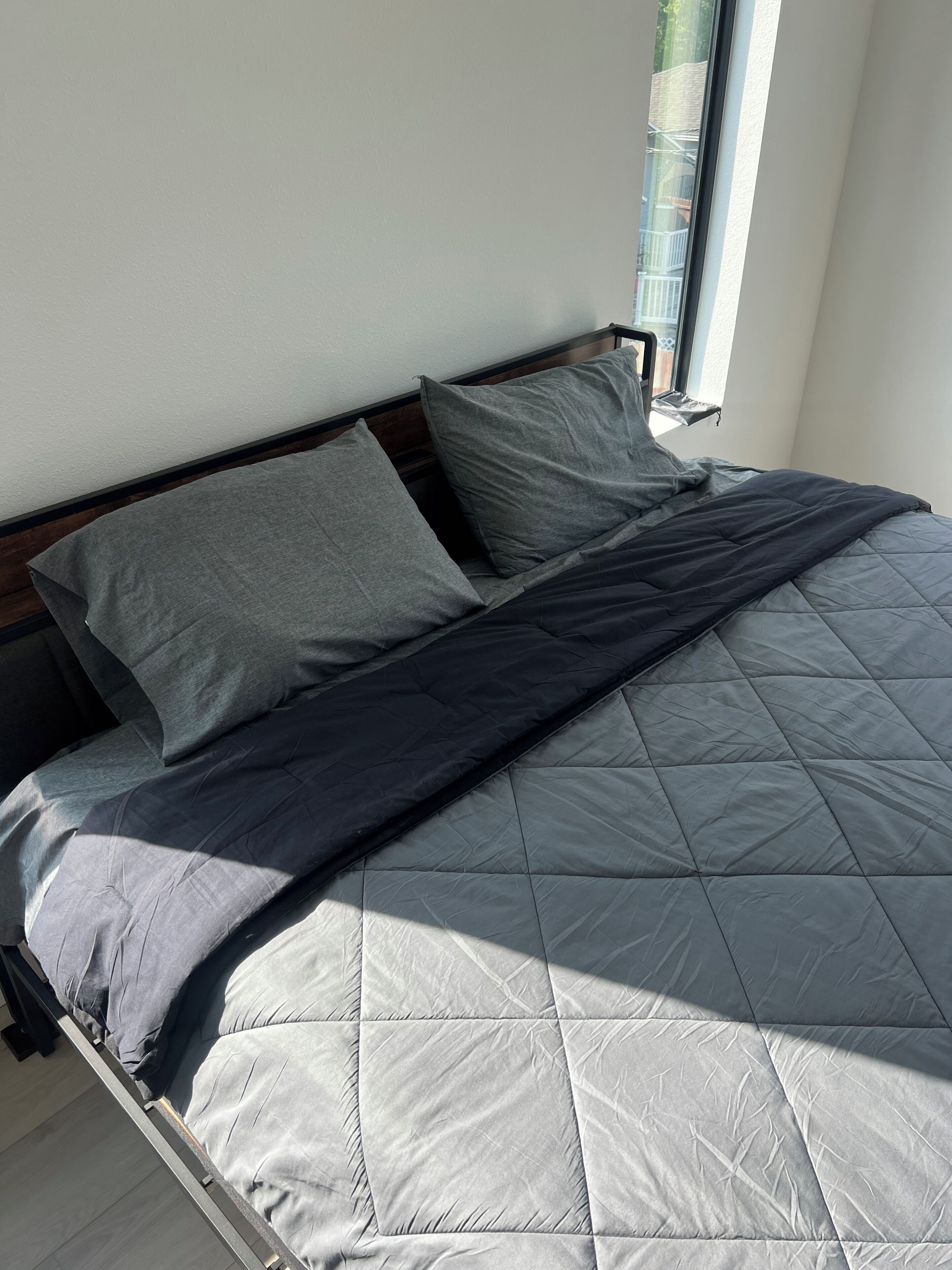 Cama com edredom cinza acolchoado, duas almofadas e cabeceira de madeira.