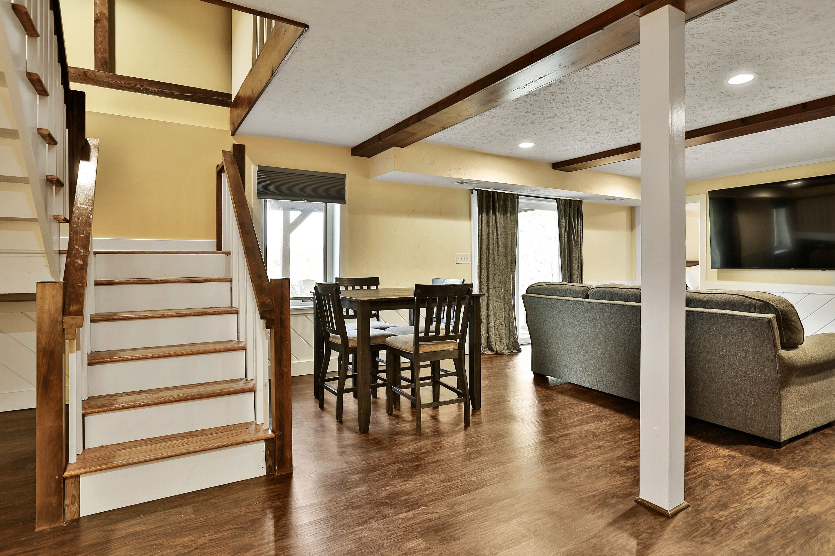 A spacious living area with a wooden staircase, a dining table with chairs, a sofa, and a flat-screen TV.