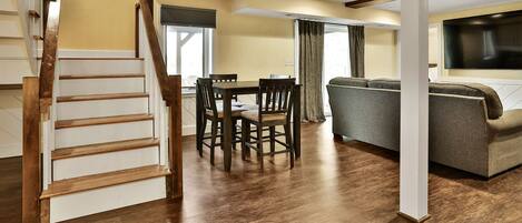 A spacious living area with a wooden staircase, a dining table with chairs, a sofa, and a flat-screen TV.