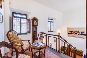Uma escadaria com corrimão de madeira, uma cadeira de madeira com almofada, uma mesa de madeira com um livro e um relógio fixado na parede.