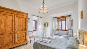 A bedroom with a large wooden wardrobe, a bed with a striped bedspread, a chair, and a nightstand with a lamp. - Chianti Classico Home Panoramic View 4, Castellina in Chianti, Italy (Castellina in Chianti)