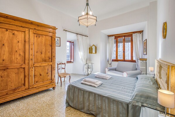 A bedroom with a large wooden wardrobe, a bed with a striped bedspread, a chair, and a nightstand with a lamp. - Chianti Classico Home Panoramic View 6, Castellina in Chianti, Italy (Castellina in Chianti)