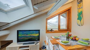 Un coin repas avec une table dressée pour deux personnes, un téléviseur posé sur un meuble et une fenêtre donnant sur l’extérieur.