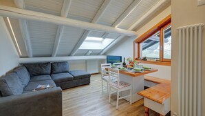 Une pièce de style « loft », avec un plafond en pente, un canapé gris, une table à manger en bois avec des chaises, et une fenêtre offrant une vue.