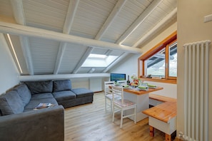 A loft-style room with a sloped ceiling, a gray sofa, a wooden dining table, and a view through a window.