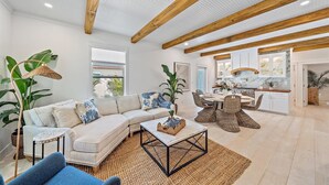 A spacious living room with a large sofa, a coffee table, and wicker chairs. There is a kitchen area with white cabinets and a dining table. - 🌸Posh Bungalow 🌴near Anna Maria Beaches, IMG and LECOM (Bradenton)