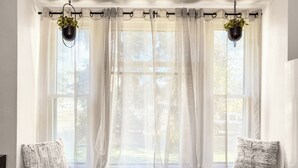 Asiento junto a la ventana con cortinas translúcidas, dos macetas colgantes y dos almohadones con textura.