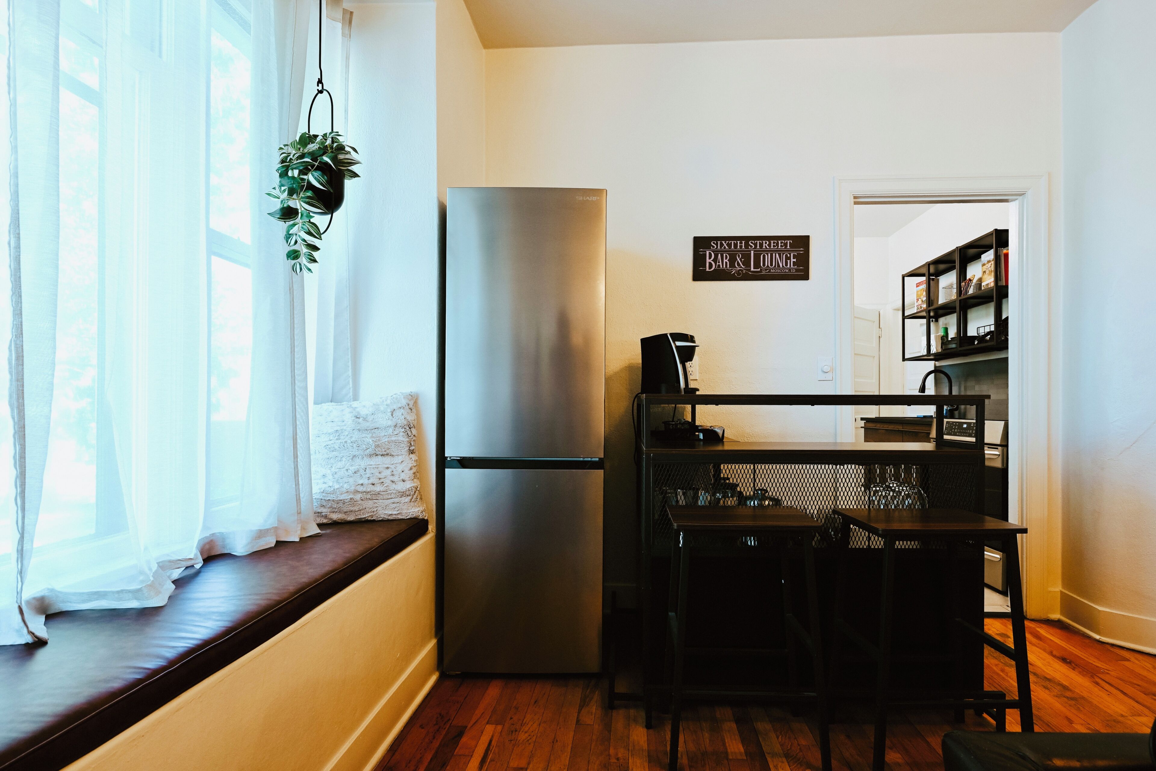 Une cuisine équipée d’un réfrigérateur encastré, une table à manger avec des chaises, une plante suspendue et une fenêtre ornée de rideaux légers.