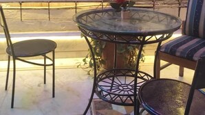 A round glass table with a potted plant and two chairs, a cushioned bench, and a view of a street at night. - Hotel Manglam Inn (Lucknow)