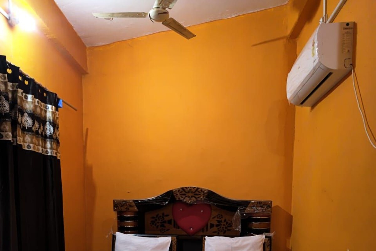 A single bed with a wooden headboard and a heart-shaped decoration, a white air conditioner on the wall, and a ceiling fan.