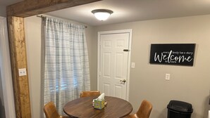 A dining area with a round wooden table, four brown leather chairs, a tissue box, and a black trash can. A white door and a 'Welcome' sign are visible in the background. - Livie’s Cozy Cottage Near Collierville (Olive Branch)