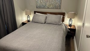 A neatly made bed with a grey bedspread and two pillows, flanked by nightstands with lamps. A painting of tall grasses hangs above the bed. - Livie’s Cozy Cottage Near Collierville (Olive Branch)