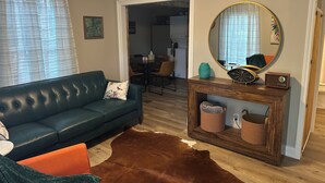 A living room with a green sofa, a brown rug, a wooden console table with a mirror and a clock, and a dining area in the background. - Livie’s Cozy Cottage Near Collierville (Olive Branch)