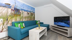 A modern living room with a blue sofa, a coffee table with a vase of flowers, a flat-screen TV on a wooden stand, and a large beach mural on the wall. - Helle Ferienwohnung fur Zwei Personen in Bensersiel (Esens)