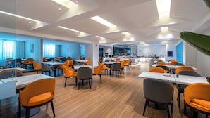A modern dining area with multiple tables and chairs, a kitchen in the background, and a bright, open ceiling design. - LiangZhi Huipin Hotel (Haikou)